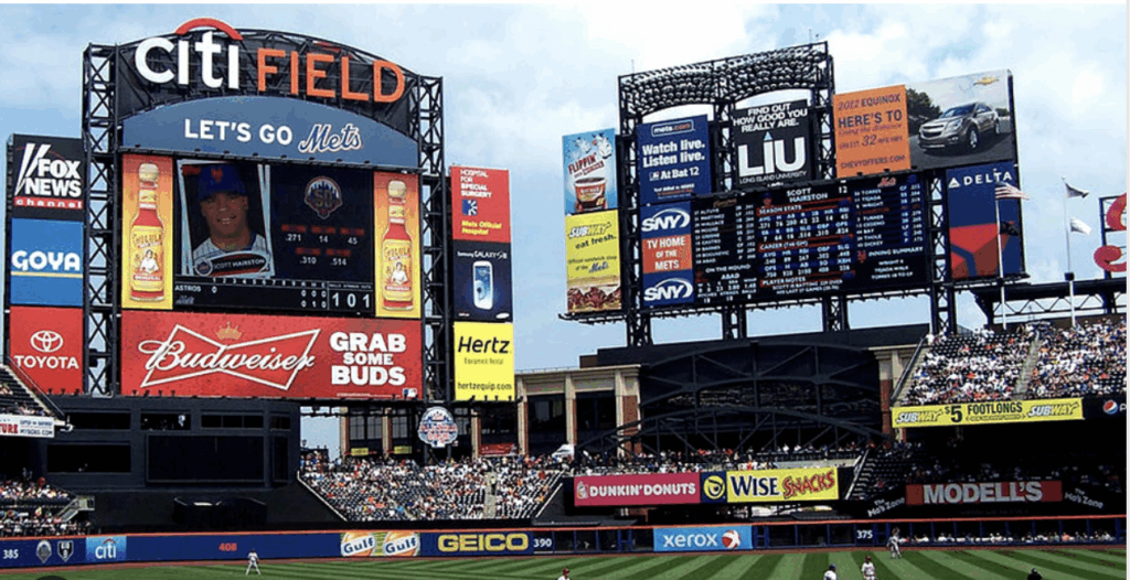 Scoreboard at Citi Field, home of the New York Mets, with crowd in stands and advertisements visible during a baseball game.