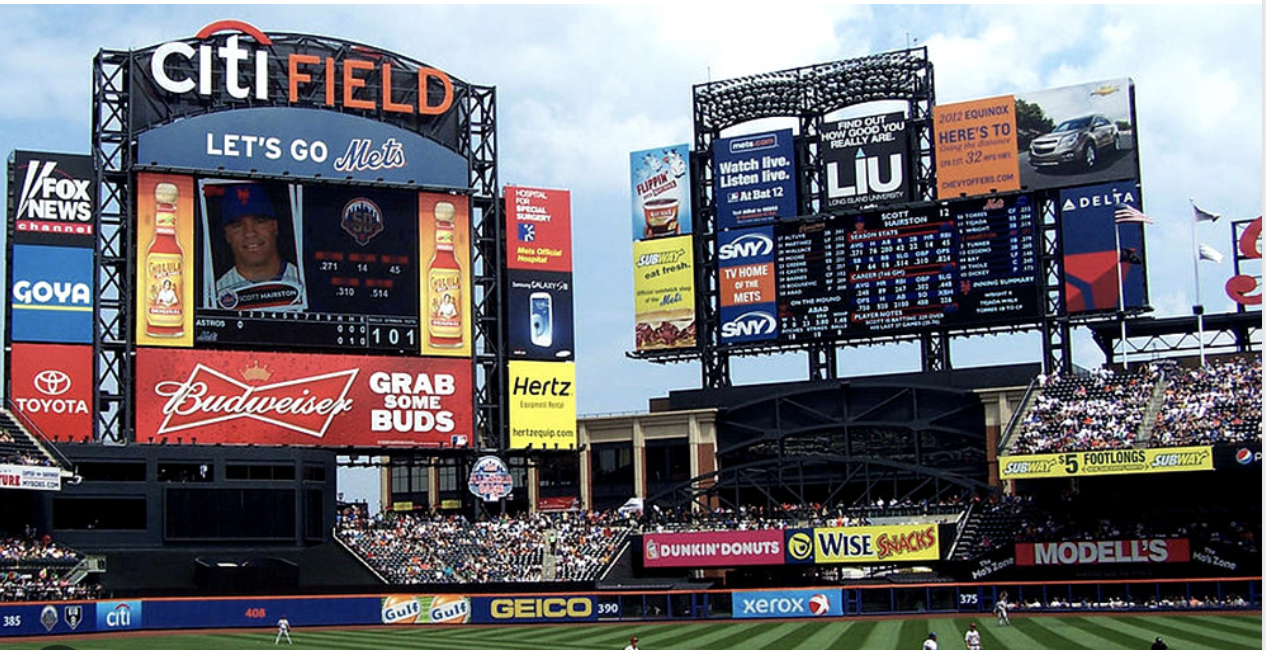 Scoreboard at Citi Field, home of the New York Mets, with crowd in stands and advertisements visible during a baseball game.